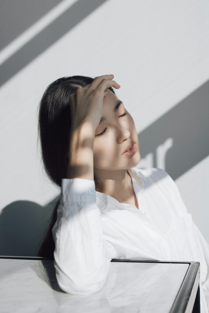 Asian woman in soft sunlight with hand to her forehead, emotion-focused AEDP therapy