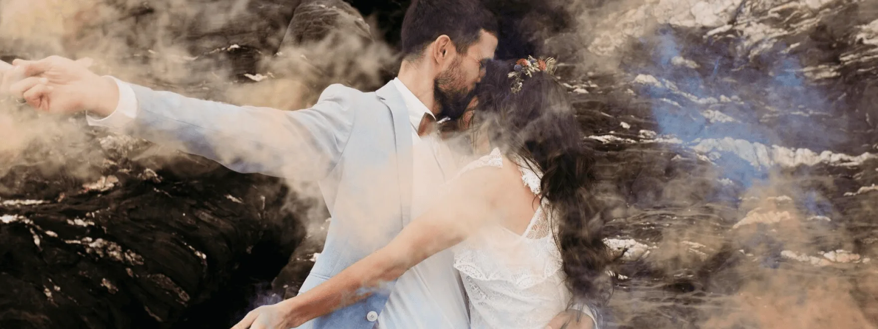 A couple in wedding attire embracing on a rocky shore, softened by drifting mist