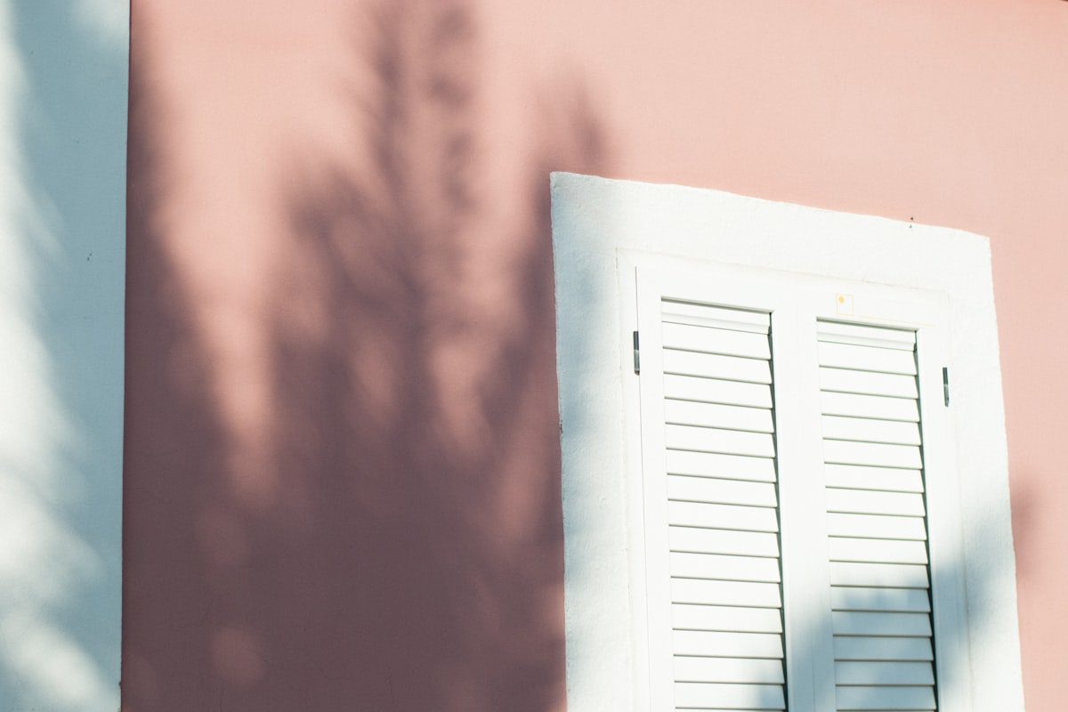 Closed white shutters on a pink wall with tree shadows