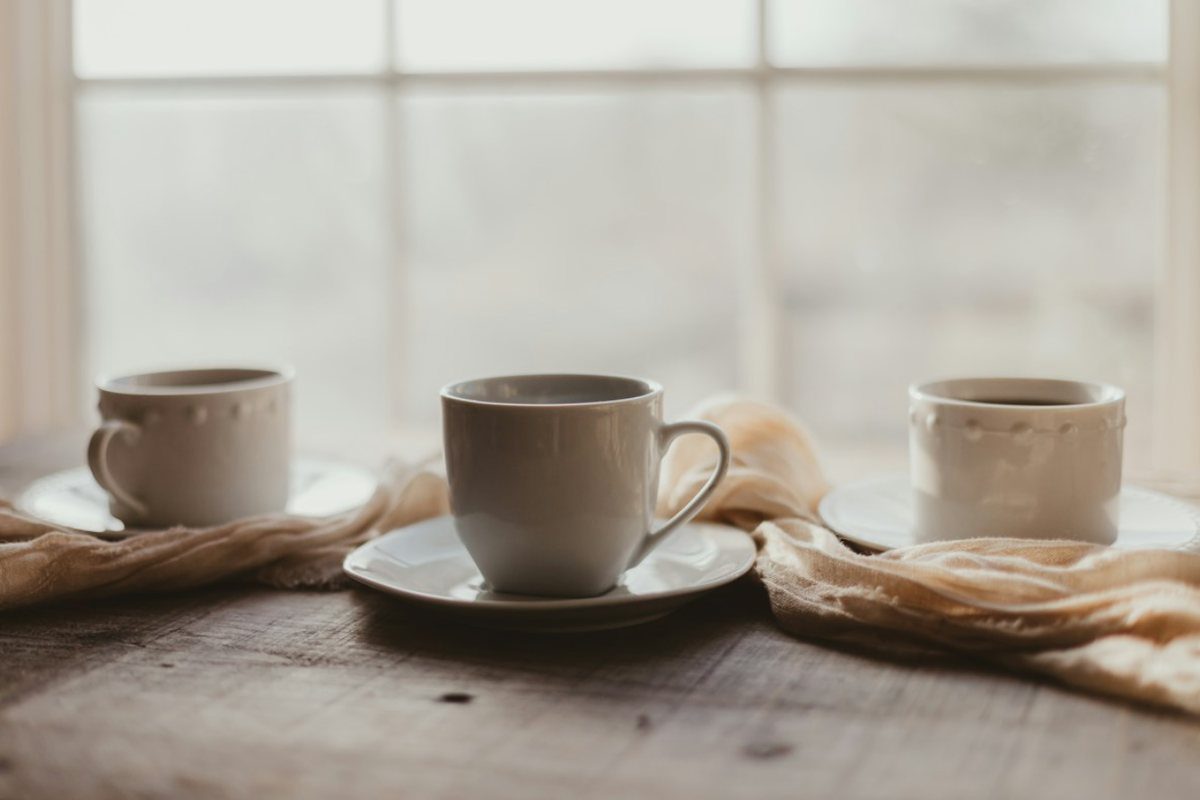 Three white cups in a row on a wooden table by a softly lit window